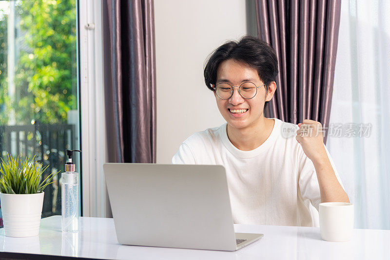 快乐亚洲年轻商业帅哥微笑在家办公戴眼镜，t恤舒适激动喜讯成功者举手是的手势与笔记本电脑在家里的桌子上