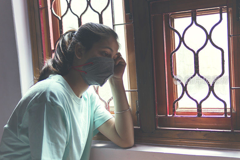 这个悲伤的女孩正在保护自己，戴着口罩，在家里自我隔离。