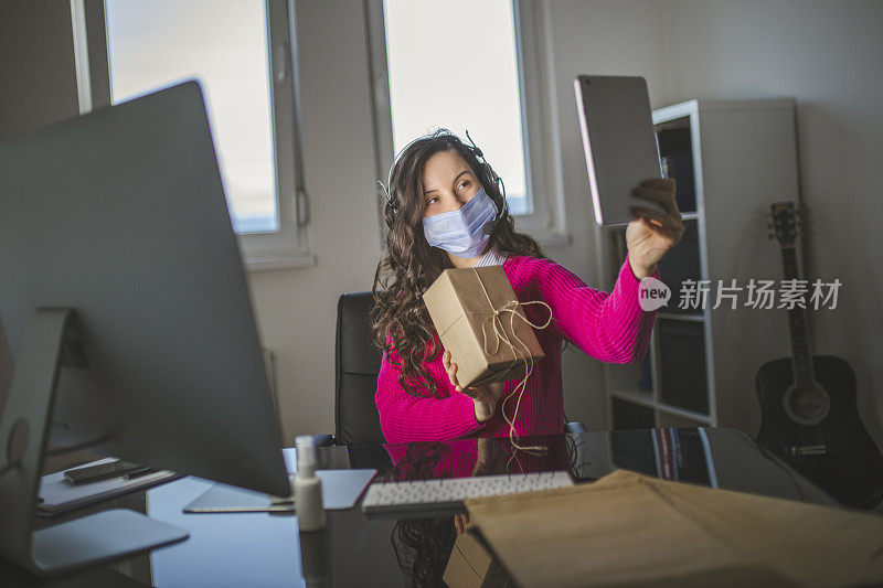 年轻女性戴着防护口罩，在平板电脑上视频通话，推广新产品，在网店工作