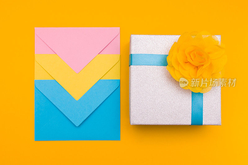 黄、蓝、粉三种颜色嵌套在一起的三个信封放在一个黄色的背景上，旁边是一个闪闪发光的礼品盒，上面有一朵黄色的花和蓝色的夏季文案空间俯视图