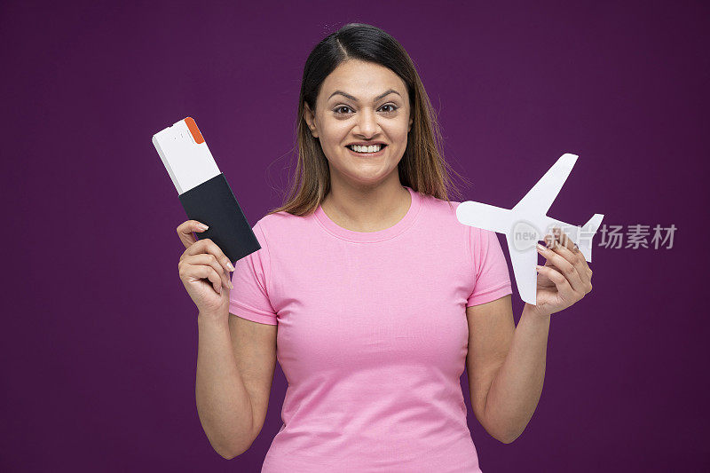 年轻女子因旅行而兴奋，拿着纸飞机，展示着机票和护照，孤立地站在紫色的背景上:-库存照片