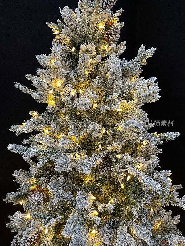 这是一棵人造云杉圣诞树，上面的塑料针上喷着人造雪，上面装饰着松果和白色的彩灯，背景是黑色的