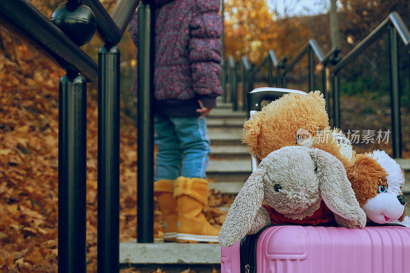 女孩站在水泥台阶上，旁边是一个装满软玩具的粉红色行李箱，走着，走着，走着，走着，走着，走着，走着，走着，走着