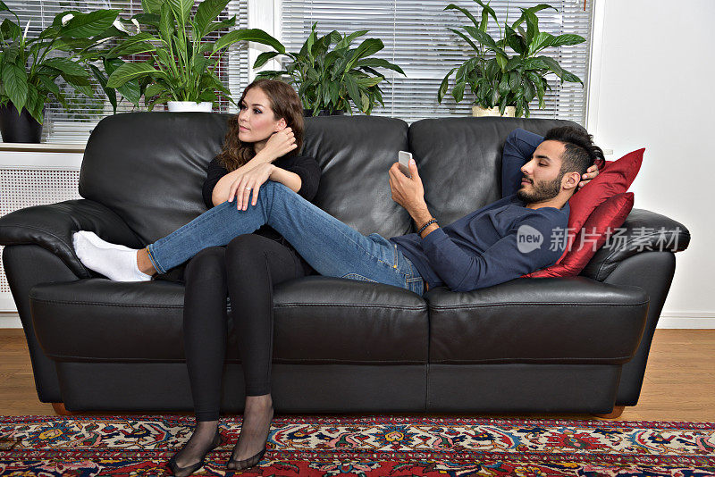 一对情侣在沙发上休息，使用手机和电视遥控器