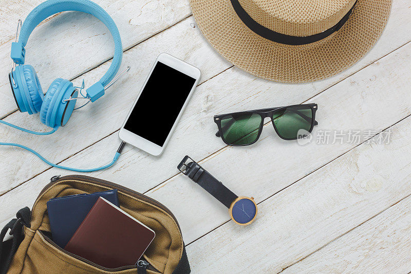 俯视图配件旅行概念。白色手机，木质背景，耳机听音乐，旅行包，护照，手表，太阳镜，帽子等必需品放在白色木质桌子上。