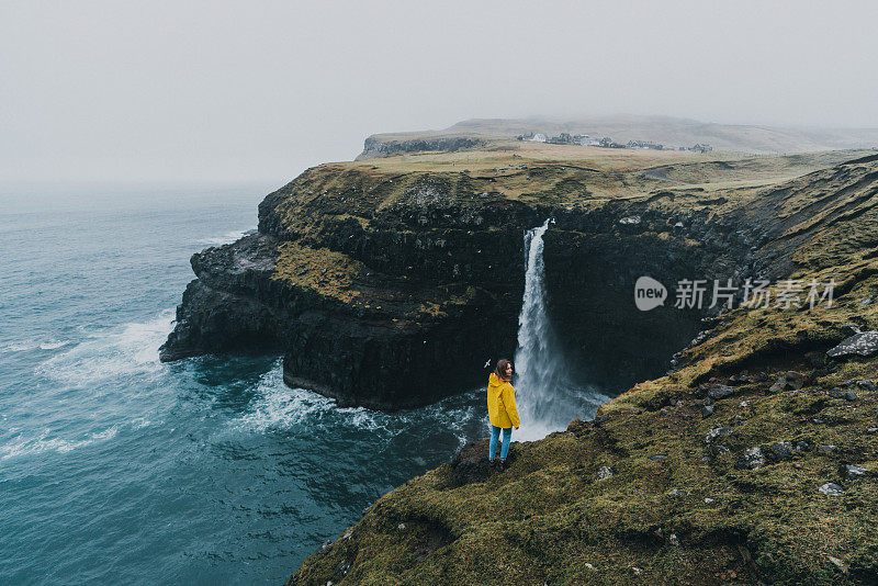 穿着黄色雨衣的女人在看风景Múlafossur瀑布掉进海里在法罗群岛