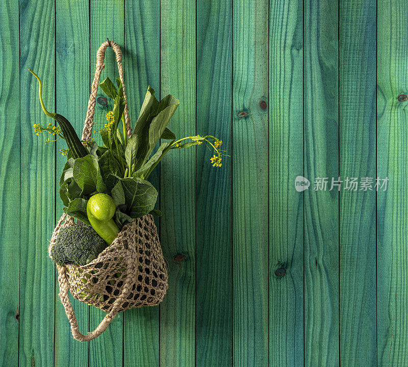 市场新鲜，绿叶蔬菜在一个天然麻绳可重复使用的购物袋挂在钩子上，以现代绿松石色木制面板背景墙。
