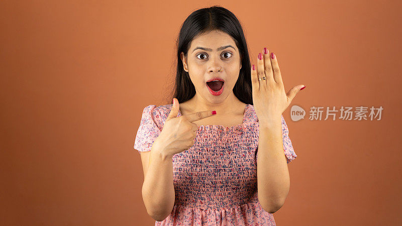 美丽兴奋的年轻印度女子穿着裙子站在孤立的彩色背景，展示她的订婚戒指在她的手指
