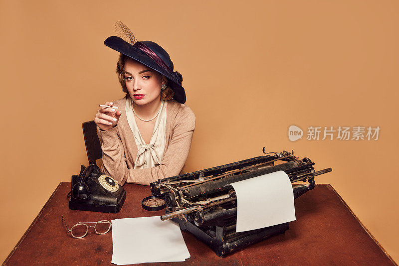 迷人的女作家穿着老式的衣服坐在桌子上，在米色的背景下吸烟