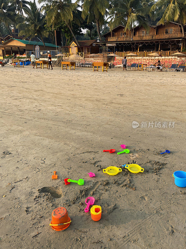 图像的塑料桶，铲子，耙子，筛子，沙模和喷壶在沙滩上，明亮的颜色的儿童海滩玩具，背景的一排海滩小屋，重点在前景