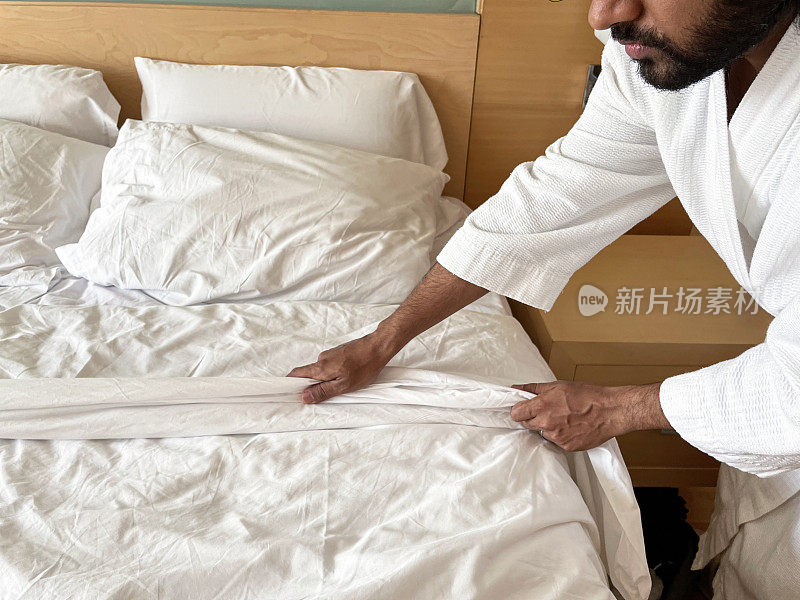 特写图像的印度男子穿着白色毛巾浴袍制作酒店双人床与木材和玻璃床头板，白色被褥被推翻，堆枕头，重点在前景