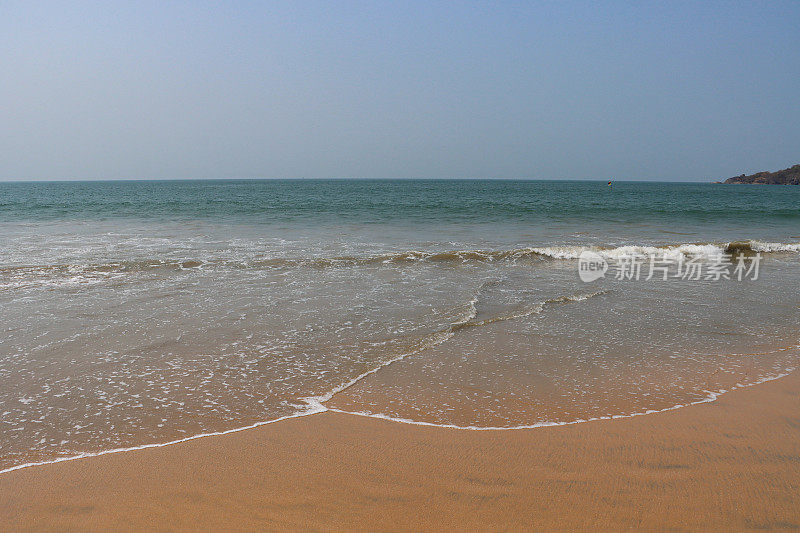 印度果阿邦Palolem海滩的海岸线，海浪拍打着海滩，树木繁茂的岛屿在地平线上，湛蓝晴朗的天空
