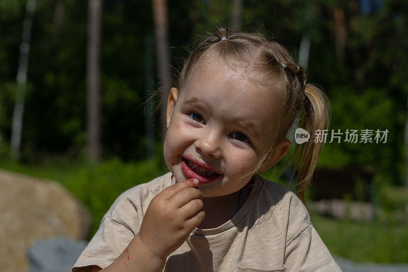 特写肖像。可爱的孩子穿着脏兮兮的草莓，微笑着看着镜头。夏天的一天，一个小女孩在吃草莓。夏天脏孩子的概念。