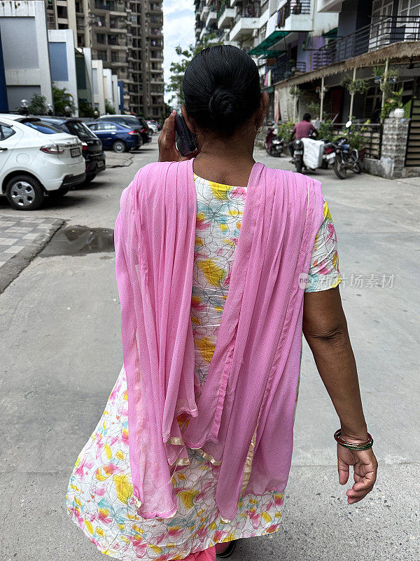 特写照片中，一名印度妇女身穿色彩鲜艳的粉色围巾和萨尔瓦·卡米兹传统服装，穿过居民区，用智能手机打电话，这是后视图，焦点是前景