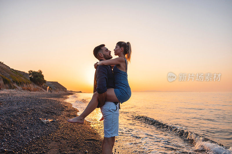 在日落时分的海滩约会中，男友将女友抱在空中，而女友则将双腿绕在他身边