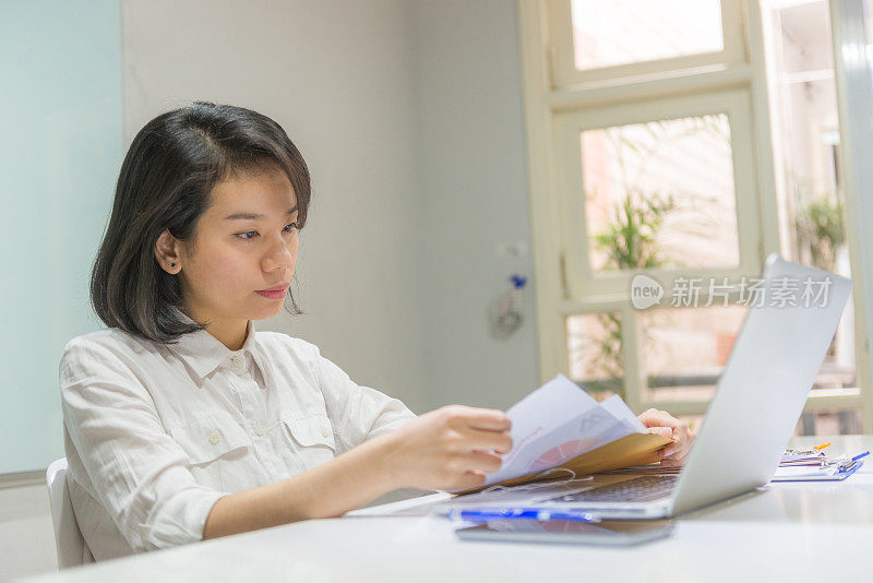 一个亚洲女人在他的办公桌上拿着文件和笔记本电脑工作