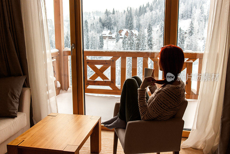 一个女人坐在舒适的椅子上，喝着咖啡，透过窗户看着白雪覆盖的山。