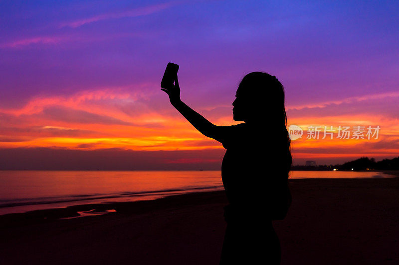 剪影的女人拍照日落与手机，在海边