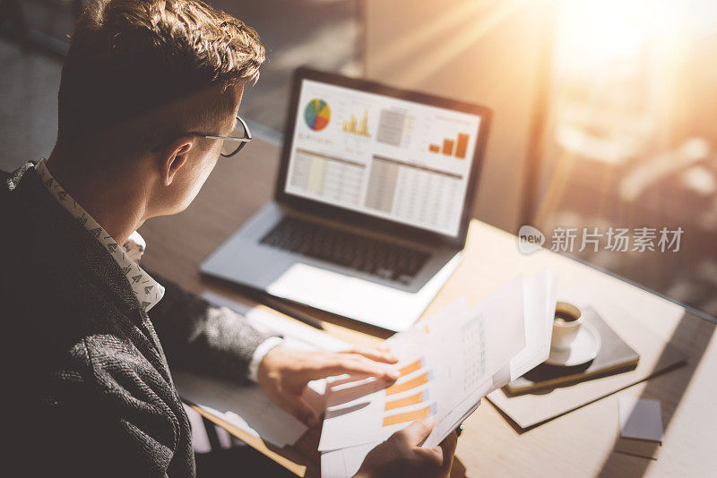 年轻的金融市场分析师戴着眼镜，坐在阳光明媚的办公室里，用笔记本电脑工作。商人分析手中的文件。笔记本屏幕上的图形和图表。