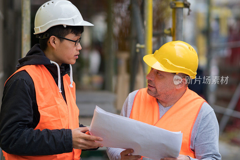 年轻的亚洲学徒在建筑工地与资深工程师工作。在户外