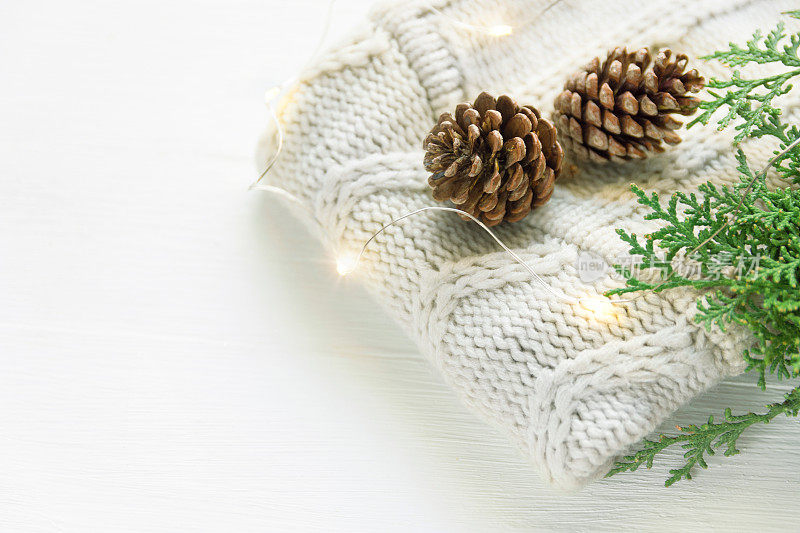 折叠的白色针织羊毛衫，小松果，绿色的杜松枝，金色的灯花环，木板木桌靠窗。舒适的冬天的晚上。新年圣诞魔幻气氛。Scandi风格