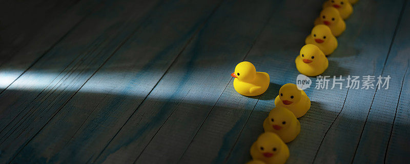 一排黄色的橡胶鸭子排成正式的线，一只鸭子脱离了线，朝着一束穿过黑暗的光线前进，场景设置在一个古老的蓝白风化的木板背景上，概念上代表水。