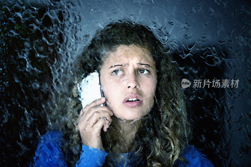 失望的年轻女子在湿窗后用智能手机聊天