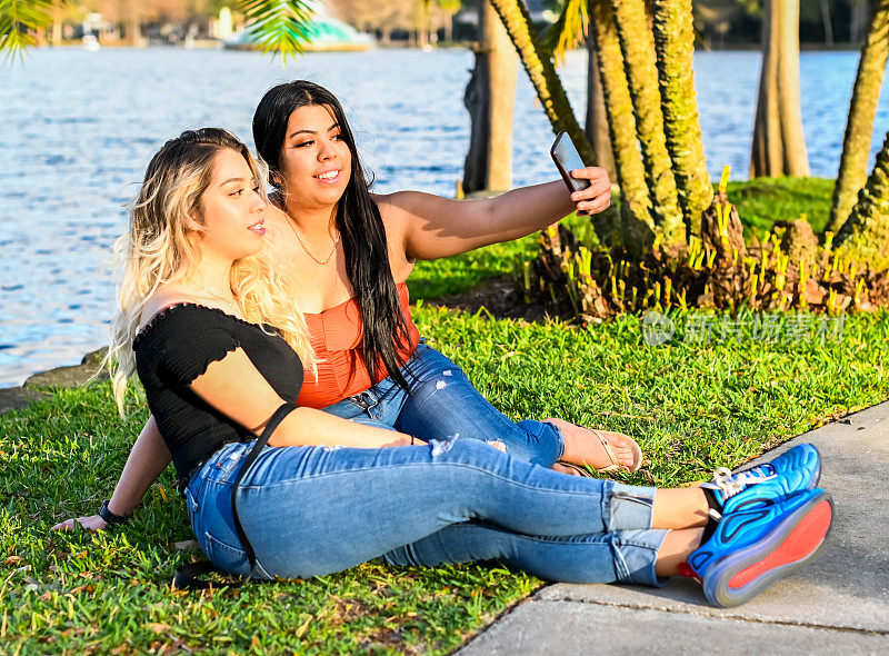 两个年轻的拉丁妇女坐在草地上拿着手机