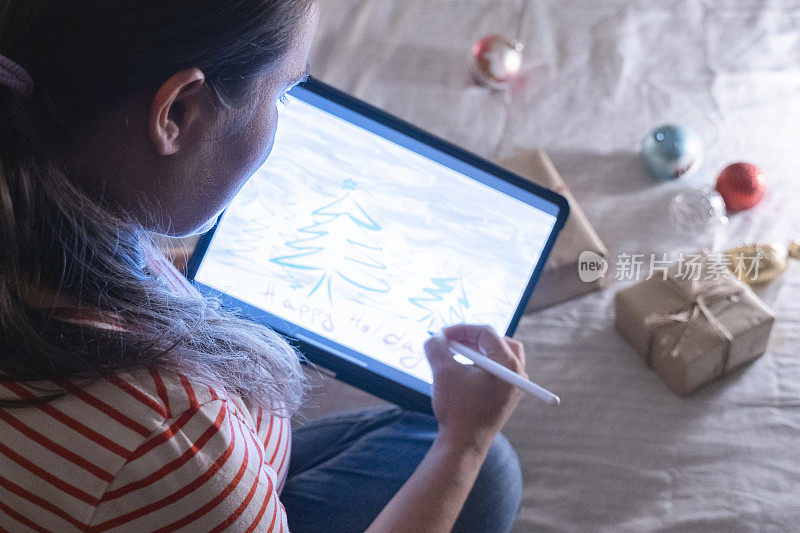 一名女性艺术家用触控笔在数码平板电脑上创作圣诞贺卡，周围是包装好的圣诞礼物。在家里讨论新冠肺炎的现实。新的标准。呆在家里准备寒假。