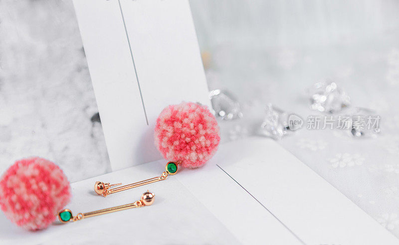 绿色宝石耳环，耳环上有粉色和白色的毛绒球。耳环旁边有透明的冰块和其他装饰。祖母绿耳环被放在铭心卡片上。这些组合看起来既优雅又时尚。