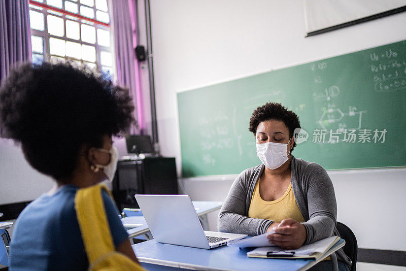 一名十几岁的学生戴着口罩在教室里与老师交谈