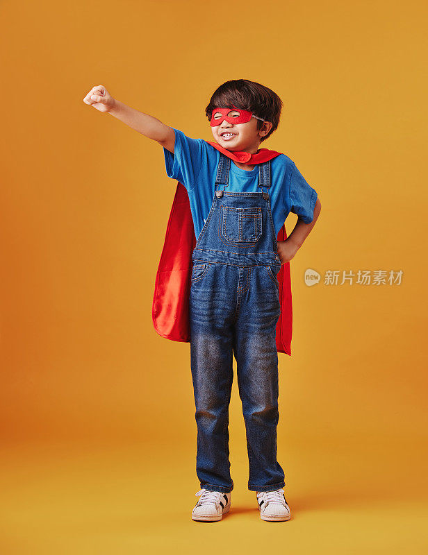 可爱的亚洲小男孩戴着面具，披着斗篷，在工作室的橙色背景下假装是超级英雄。可爱快乐的男孩为了万圣节假装成一个角色