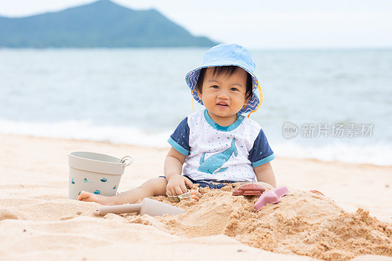 可爱的婴儿蹒跚学步坐在沙滩上玩耍和学习。第一次触摸沙子的可爱宝宝宝宝兴奋和幸福。发展宝宝的技能和经验。健康健康的生活