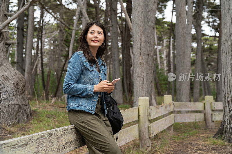 微笑的亚洲中国女旅行者坐在低矮的篱笆上放松，享受自然美景，参观克罗克树林沿着17英里的车程在美国加利福尼亚州