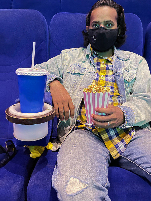印度男子戴着口罩，坐在一排电影院座位上，拿着焦糖爆米花的纸板盒，一次性饮料杯和饮料架中的吸管，关注前景