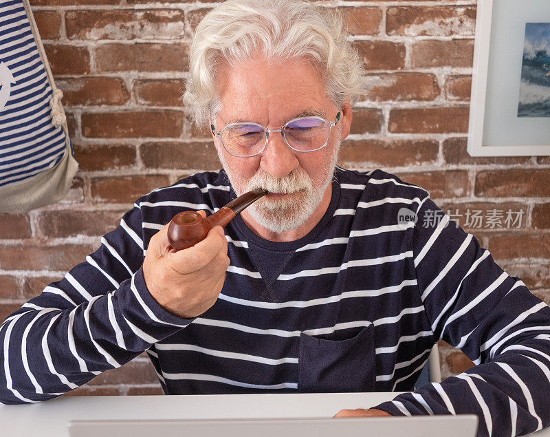 老人在家里用手提电脑抽烟斗。砖墙作为背景和装饰