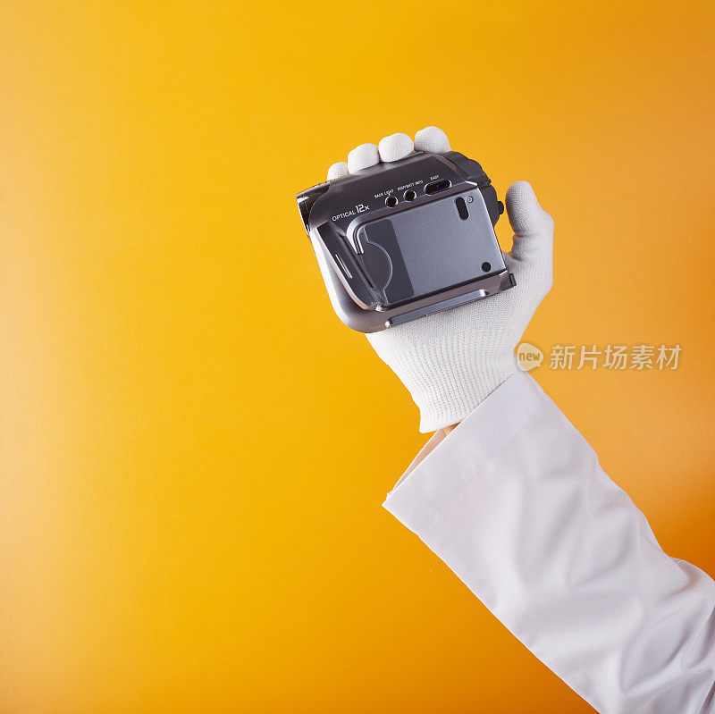 技术员戴着白色手套，手持迷你DV摄像机，背景为黄橙色