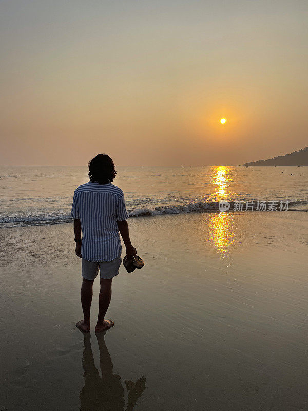 在印度果阿的帕洛勒姆，在太阳开始落山的黄昏，在果阿海滩度假时，一个无法辨认的人在海边划桨，度假的游客光着脚走在海滩上，手里拿着鞋子在水里戏水，后景，聚焦于前景