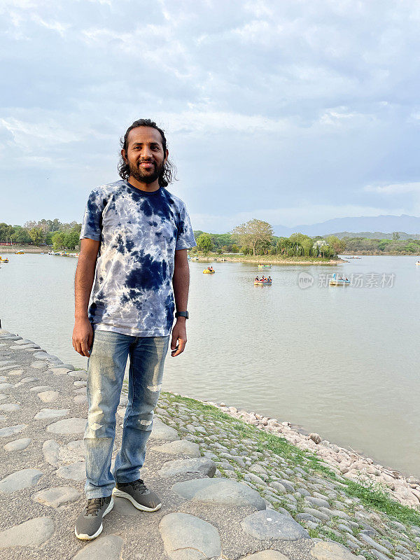 印度男子在印度昌迪加尔苏赫纳湖岸的特写照片，他看着照相机、水库水面上的船只、独木舟、脚踏和皮划艇、候鸟保护区、国家湿地