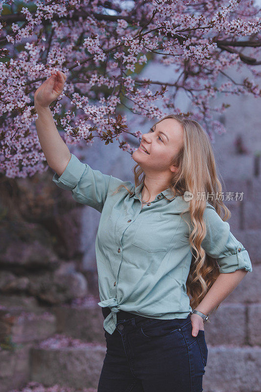 美丽的年轻女子正在享受春天，她站在一棵盛开的树旁，抚摸着粉红色的花朵