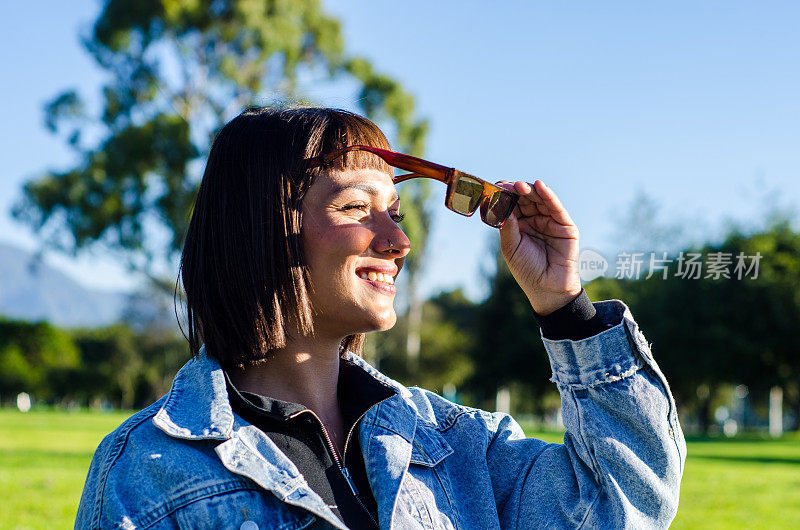 在一个阳光明媚的日子里，一名剪短头发的年轻女子在公园里穿着休闲服，戴着太阳镜