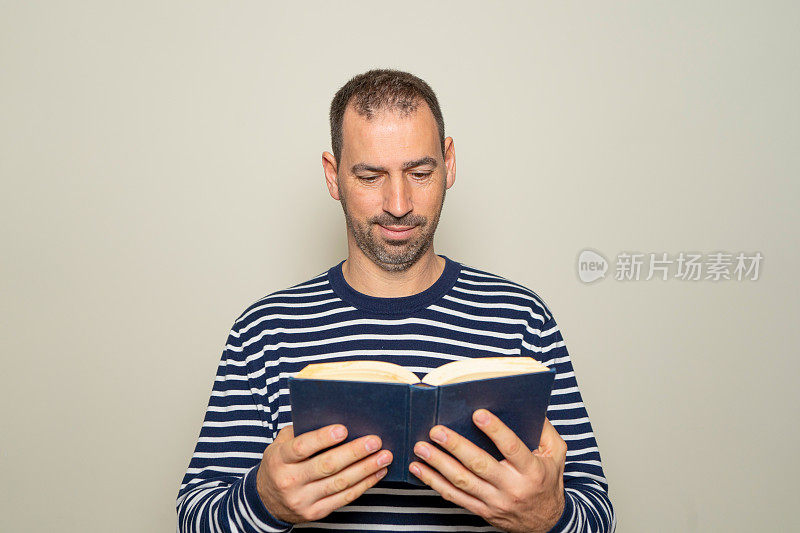 40多岁的西班牙裔男子留着胡子，穿着条纹毛衣，正在读一本纸质书，他对故事的精彩内容感到专注和兴奋。孤立的米色背景。