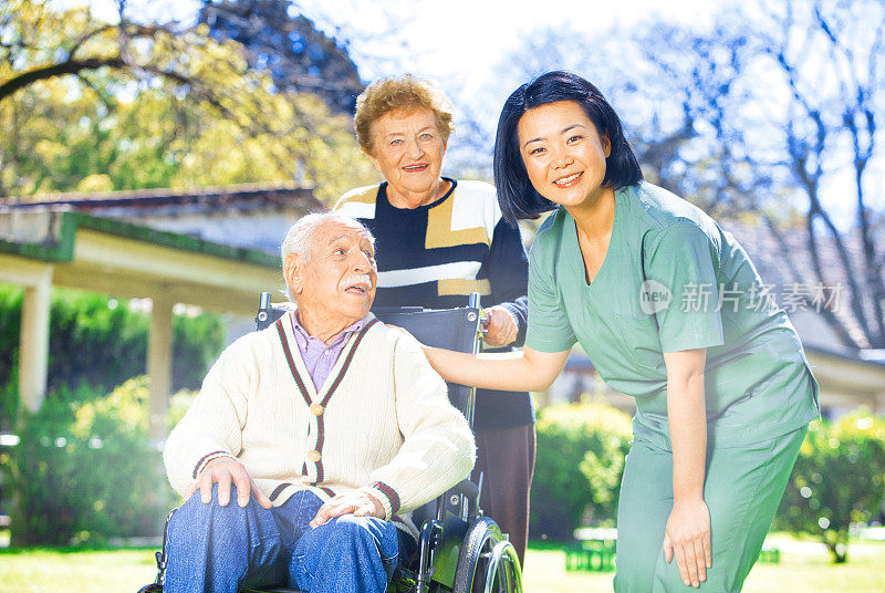 残疾。年长的女士与丈夫在轮椅由亚洲护士协助花时间在一个阳光明媚的早晨花园。辅助生活。