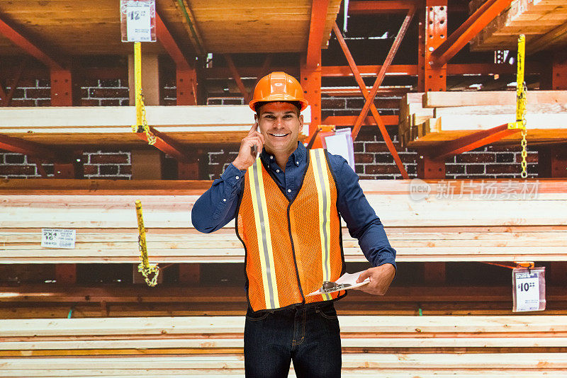 年轻的男性建筑工人站在仓库的商品前，穿着反光的衣服，拿着剪贴板，使用智能手机