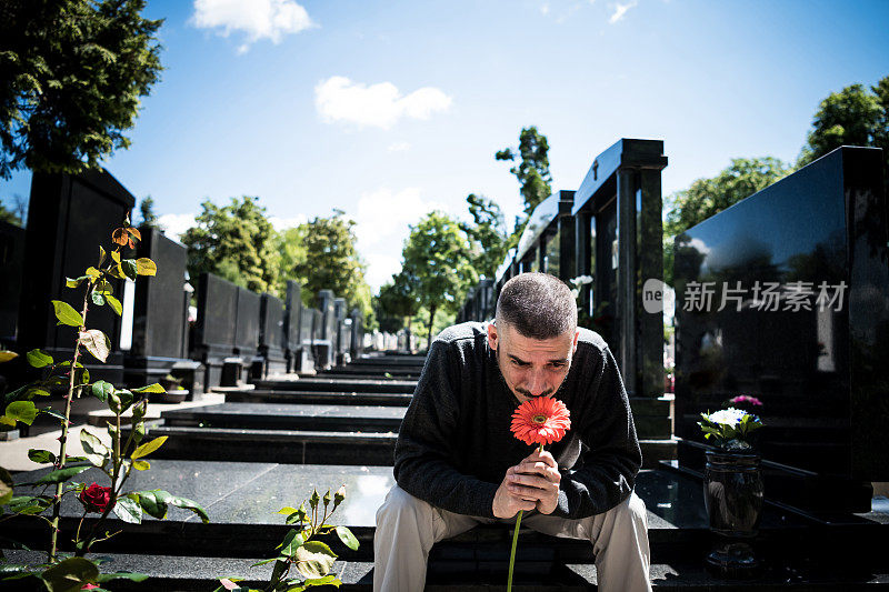 悲伤和沮丧的鳏夫在黑衣跪在墓碑前，拿着一朵花和哀悼家人的损失。死亡、哀悼、葬礼和精神的概念——Covid-19