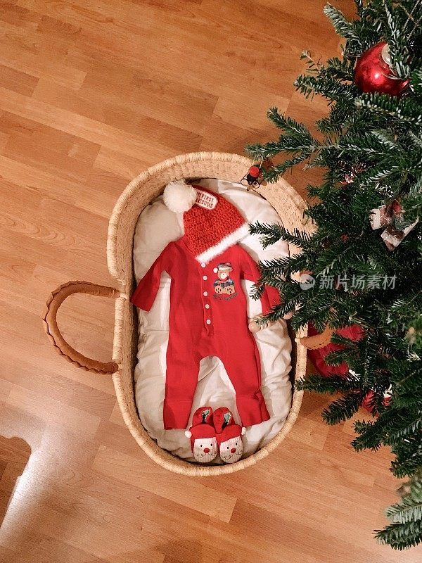 手工制作的摩西篮子放在真正的圣诞树下，圣诞树上装满了圣诞婴儿服装，等待着一个圣诞男婴