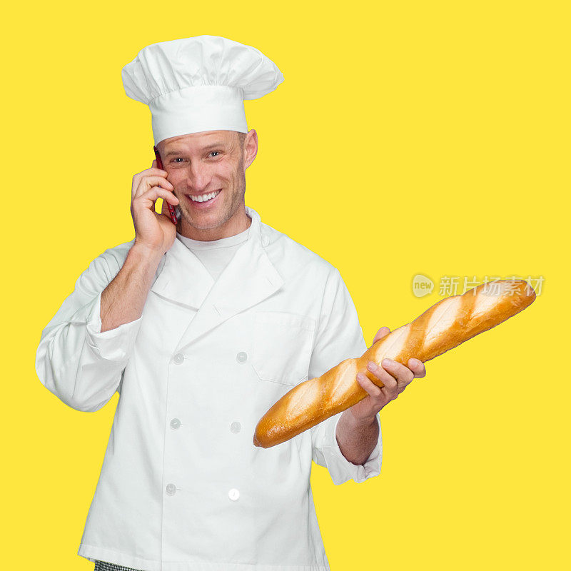 白人男性面包师穿着制服，拿着法棍面包，用智能手机站在黄色背景前