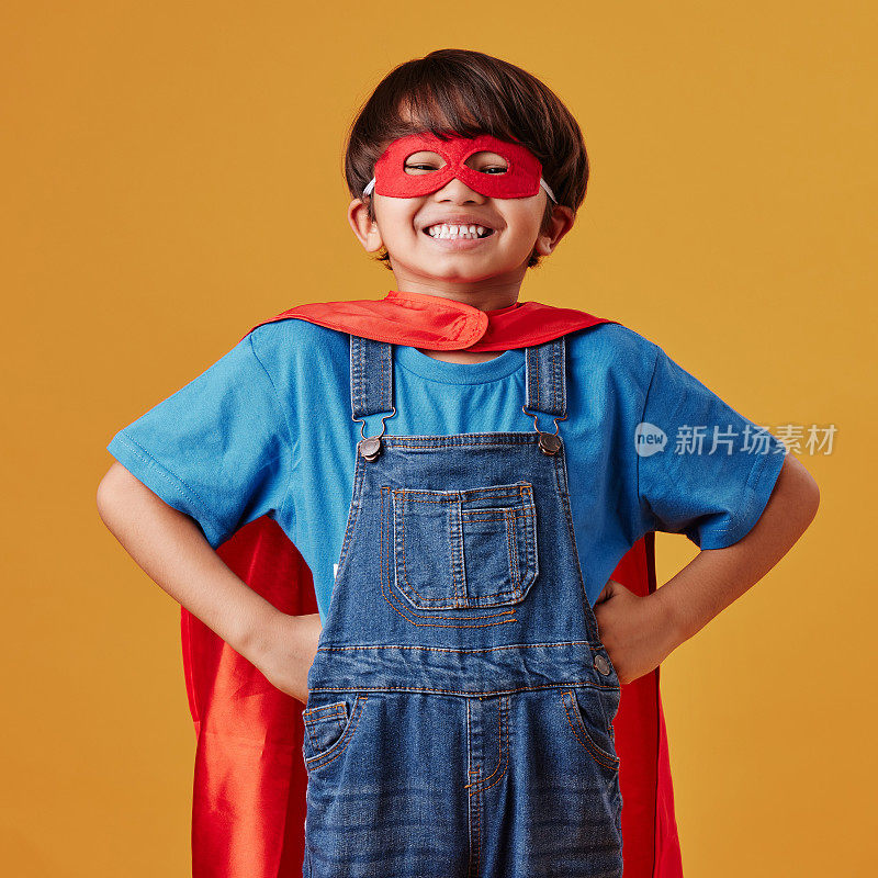 可爱的亚洲小男孩戴着面具，披着斗篷，在工作室的橙色背景下假装是超级英雄。可爱快乐的男孩为了万圣节假装成一个角色
