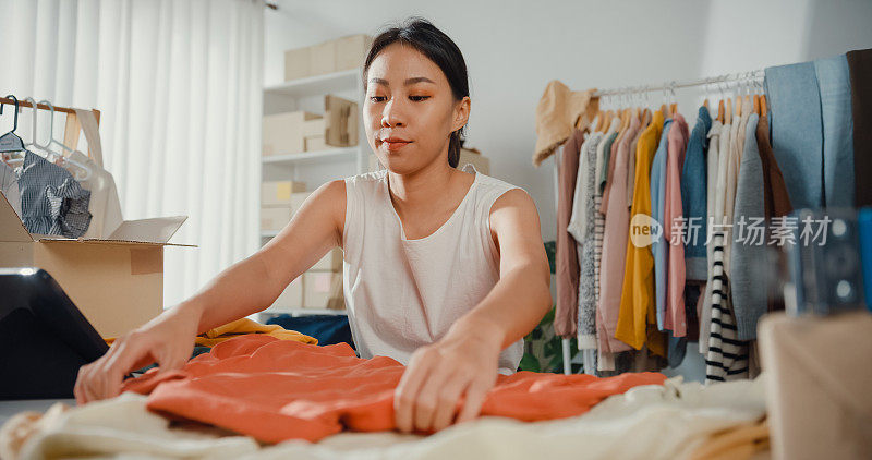年轻的亚洲小企业主准备包装订单衬衫产品发送客户与库存检查平板电脑在家里。
