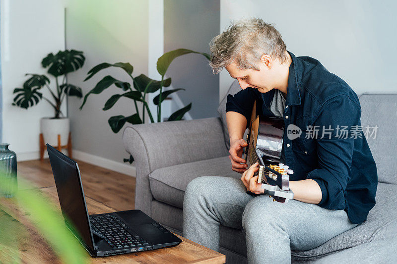 一名男子正在家里用笔记本电脑上在线吉他课，坐在客厅的灰色沙发上。他在电脑上看原声吉他教程。新的爱好。有选择性的重点。